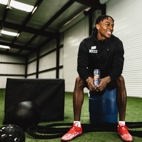 Man sitting on a gym roller holding a drink can, smiling in a gym.